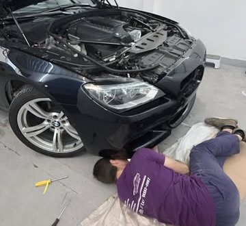Marcelo deitado no chão fazendo reparo sob o capô aberto de um BMW